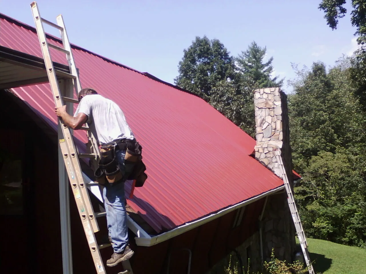 Roofer on a ladder beside a red metal roof and stone chimney — Soffit & Fascia Repair by Roofing Brothers in Keansburg